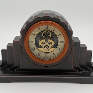 May include: A dark brown mantel clock with a stepped, tiered base and a circular clock face. The face features a gold-toned background, black Roman numerals, and a visible mechanical movement in the center, surrounded by a smooth, circular frame.