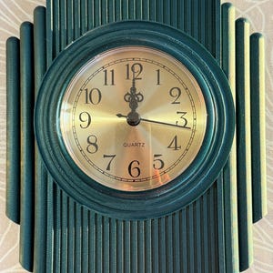 May include: A teal-colored quartz wall clock with a gold-colored face. The clock has a round face with black numerals and hands. The clock's frame has a vertical, ribbed design with rounded edges.