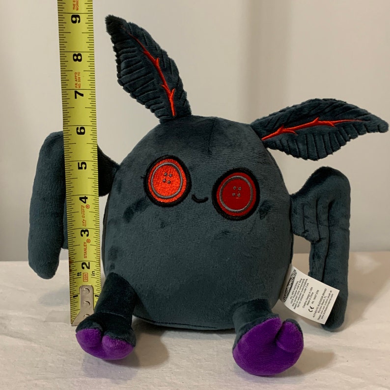 May include: A gray plush toy with large red button eyes and purple feet. The toy has two large gray wings and two smaller gray ears with red veins. The toy is sitting on a white surface.
