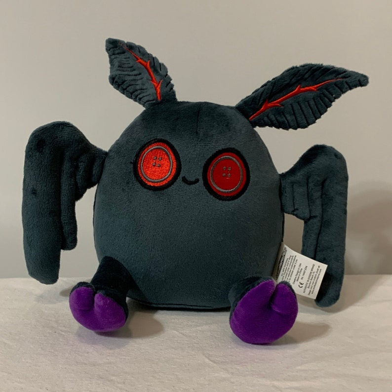 May include: A plush toy shaped like a bat with large red button eyes. The bat is gray with purple feet and black wings. The toy has a small tag on the side.