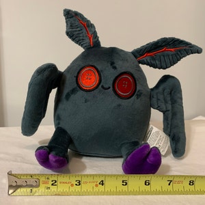 May include: A plush toy shaped like a bat with large, red button eyes. The toy is gray with purple feet and red, leaf-shaped ears. The toy is approximately 4 inches tall.