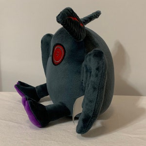 May include: A plush toy shaped like a round, gray creature with purple feet and a red eye. The creature has two large, gray wings.