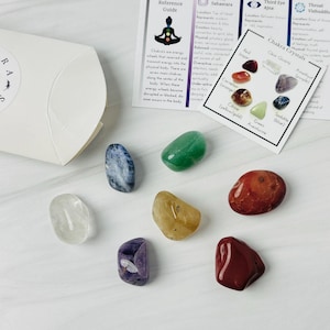 May include: Set of seven chakra crystals, including clear quartz, amethyst, sodalite, aventurine, carnelian, citrine, and red jasper. The crystals are arranged in a circle on a white surface, with a card showing the chakra colors and names.