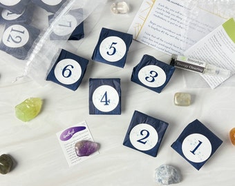 May include: An advent calendar with numbered navy blue packages, each containing a crystal. The calendar includes a smudge spray, amethyst, and other crystals. The image also shows a card with text.