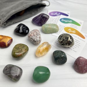 May include: A collection of polished tumbled gemstones including amethyst, amazonite, pyrite, and various other multi-colored stones. The set includes three informational cards labeled Amethyst, Amazonite, and Pyrite, and a grey fabric drawstring pouch.