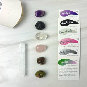 May include: A selection of polished gemstones, such as amethyst, Apache tear, clear quartz, rose quartz, smoky quartz, and unakite, are arranged with a white selenite wand and information cards. The cards detail each stone's name and properties.