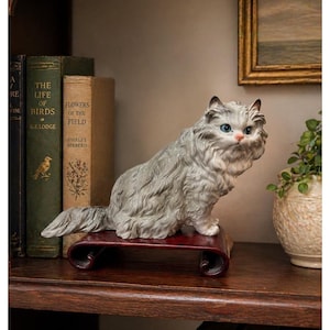 May include: A ceramic figurine of a gray cat with blue eyes, perched on a dark red wooden stand. The cat has fluffy fur and is posed in a seated position. The figurine is displayed on a wooden shelf with antique books.