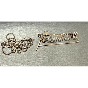 May include: Two vintage gold-toned wire name pins. One pin spells out "Stacey" in a cursive font. The other pin spells out "Lorraine" in a similar cursive style. Both pins are displayed on a grey surface.