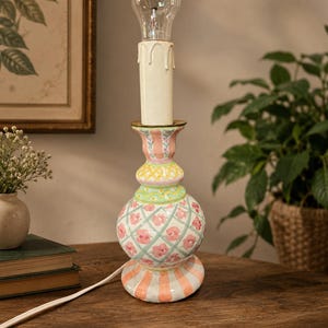 May include: A decorative ceramic table lamp with a bulb. The lamp has a cream-colored candle-shaped top, a bulb, and a base with pink, orange, and green floral patterns. The lamp is on a wooden surface.