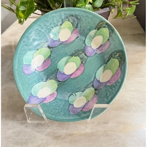 May include: A turquoise ceramic plate with a floral design. The flowers are composed of green, white, and purple petals. The plate is displayed on a clear acrylic stand. The plate has a glossy finish.