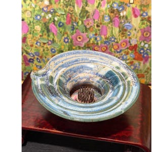 Peut inclure: Un crapaud de fleur en céramique avec une finition émaillée bleue et verte. Le crapaud a une forme circulaire avec une ouverture centrale et un porte-épingles pour arranger les fleurs. Le crapaud est exposé sur une surface en bois foncé.