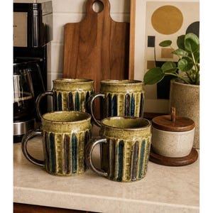 Könnte beinhalten: Vier Keramikbecher mit geripptem Design in Olivgrün, Braun und Blau. Die Becher haben dunkelbraune Henkel und stehen auf einer hellen Arbeitsplatte. Ein Holzschneidebrett und eine Kaffeemaschine befinden sich im Hintergrund.