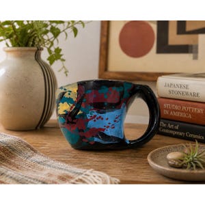 May include: A black ceramic mug with a large handle, decorated with colorful paint splatters in blue, yellow, and red. The mug sits on a wooden surface, next to a vase and books.