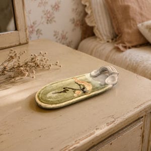 May include: An elongated oval ceramic tray featuring a glossy sage green glaze with a hand-painted abstract black line and a textured tan accent. The tray includes a sculpted, raised handle on one end with a smooth, light gray finish.
