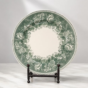 May include: A round ceramic plate featuring a cream-coloured centre and a wide border decorated with a dark green floral pattern of roses and leaves. The edge of the plate is finished with a repeating circular dot design, displayed on a black stand.