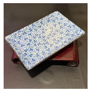 May include: Rectangular ceramic serving dish with a blue and white floral pattern. The dish has a gold rim and is displayed on a dark wooden stand. The design features intricate blue leaves and vines against a white background.
