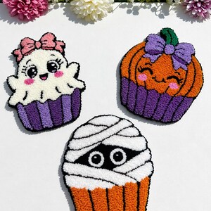 Puede incluir: Tres piezas de arte punch needle con motivos de Halloween: un fantasma blanco con lazo rosa en un molde de cupcake morado, una calabaza naranja con lazo morado en molde morado y una momia blanca con ojos visibles en un molde naranja.