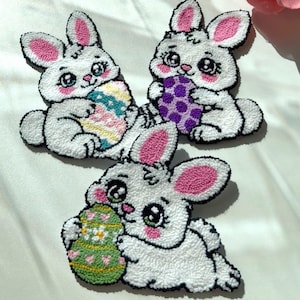 Puede incluir: Tres decoraciones de Pascua con forma de conejito blanco. Cada conejito tiene orejas internas rosas, mejillas rosadas y sostiene un huevo de Pascua colorido. Los huevos están decorados con patrones en colores pastel. Los conejitos son de material texturizado y esponjoso.