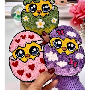 Może przedstawiać: Trzy kolorowe, ręcznie robione podstawki z kurczętami w jajkach. Podstawki z kurczętami są żółte z niebieskimi oczami i znajdują się w jajkach w kolorze zielonym z białymi kwiatami, różowym z czerwonymi sercami i fioletowym z białymi chmurami. Podstawki są trzymane przez osobę.