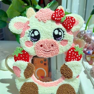 May include: A tufted wall mirror shaped like a cow with white, pink, and brown yarn. The cow features a red strawberry bow, large black eyes with white highlights, and two red strawberries held by its front hooves. The mirror is circular.