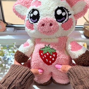 Puede incluir: Un peluche de vaca hecho a mano, en colores rosa y blanco. El juguete tiene ojos grandes y expresivos, orejas rosas y un diseño de fresa en el pecho. El juguete está hecho con la técnica de punch needle.