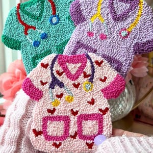May include: Three colorful, textured, handmade scrub tops. The pink scrub top has a heart-shaped collar and red heart accents. The green and purple tops also feature heart-shaped collars and stethoscope designs. These decorative items are perfect for medical professionals.