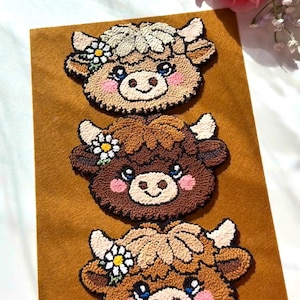 Op de afbeelding: Drie handgemaakte koeiengezichtpatches met geborduurde details. Elke patch heeft een andere bruintint, met witte accenten, blauwe ogen en een madeliefjeskroon. De patches zijn gerangschikt op een mosterdkleurige viltachtergrond.