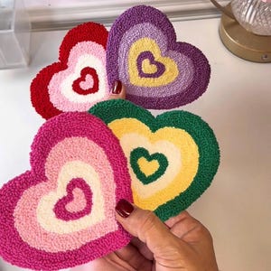 May include: A collection of heart-shaped, tufted coasters in various colors, including red, pink, purple, green, and yellow. Each coaster features a heart-shaped design in the center. The coasters are displayed on a white surface.