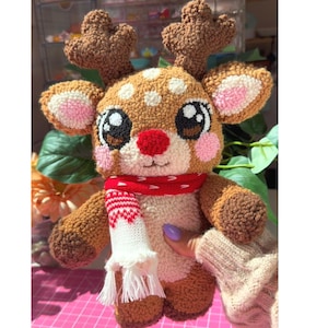 May include: A plush reindeer toy with brown and cream textured fabric, large dark eyes, a bright red nose, and pink cheeks. The reindeer wears a red and white knit scarf with a fringe detail. The antlers are brown and textured.