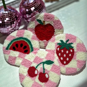 May include: Four round, handmade coasters with fruit designs. Each coaster features a pink and white checkered pattern. The fruits include a red apple, a watermelon slice, a strawberry, and cherries. Two disco ball ornaments are in the background.
