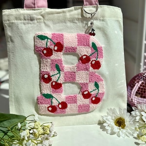 May include: A canvas tote bag with pink handles and a cherry-themed charm. The bag features a large, pink and white checkered letter "B" decorated with red cherries and green leaves. The bag is off-white.
