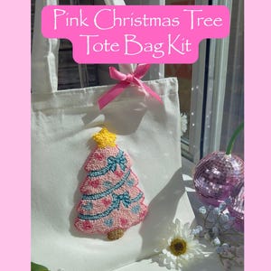 Può includere: Una borsa tote bianca con un design a forma di albero di Natale rosa. L'albero è decorato con fiocchi blu e una stella gialla. Il testo "Pink Christmas Tree Tote Bag Kit" è visualizzato su uno striscione rosa.