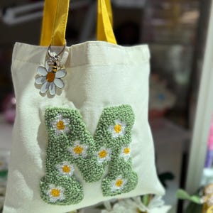 May include: Cream canvas tote bag with yellow straps. The bag displays a green, daisy-covered letter "M" and a daisy and bee charm. The bag is a light color and has a simple design.