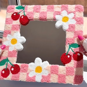 May include: A square mirror featuring a tufted, textured frame with a pink and light pink checkered pattern. The frame is decorated with three white daisies with yellow centers and two pairs of red cherries with green stems and leaves.