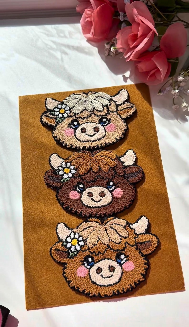 Op de afbeelding: Drie handgemaakte koeienkop-appliqu&eacute;s in bruin-, beige- en cr&egrave;metinten, elk met een madeliefje en roze wangen. De applicaties zijn gerangschikt op een bruine viltachtergrond.