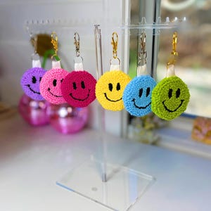 Puede incluir: Un conjunto de seis llaveros circulares de punch needle colgados en un soporte de exhibición de acrílico transparente. Cada llavero presenta una cara sonriente de distinto color: morado, rosa claro, rosa oscuro, amarillo, azul y verde, con cierres de metal dorado.