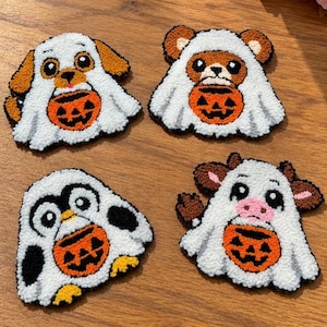 Puede incluir: Cuatro parches textiles con relieve que muestran un perro, un oso, un pingüino y una vaca, cada uno con un disfraz de fantasma blanco y sosteniendo un cubo de dulces de calabaza naranja. Diseños de hilo texturizado en blanco, naranja, negro y marrón.