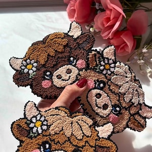 May include: Three handmade cow-shaped coasters. Each coaster features a brown, tan, and cream colour scheme with a daisy flower crown. The coasters have pink cheeks and blue eyes. The coasters are made of a textured material.