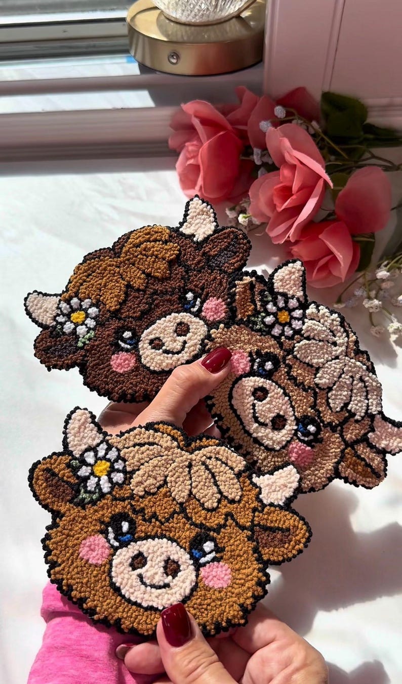 May include: Three handmade cow-shaped coasters. Each coaster features a brown, tan, and cream colour scheme with a daisy flower crown. The coasters have pink cheeks and blue eyes. The coasters are made of a textured material.