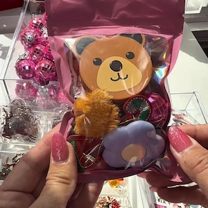 May include: A pink cellophane bag containing various decorative items. Inside are a bear-shaped ornament, a fluffy orange pom-pom, a purple flower-shaped item, and a pink, green, and silver ornament. Other items are visible in the background.