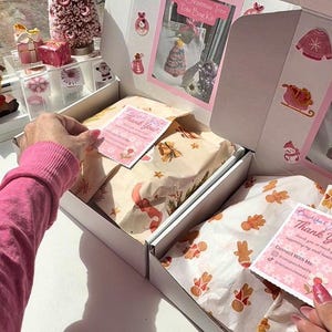 May include: A white box with pink Christmas decorations and contents. The box contains wrapped gifts and cards with "Thank You" on them. A pink Christmas tree and other decorative items are visible in the background.