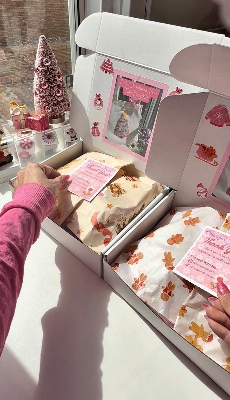 May include: A white box with pink Christmas decorations and contents. The box contains wrapped gifts and cards with "Thank You" on them. A pink Christmas tree and other decorative items are visible in the background.