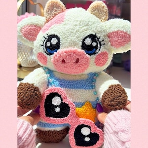 May include: A handcrafted plush cow toy with a white body, pink and brown details, and large, expressive eyes. The cow is dressed in a blue and white striped outfit and holds pink heart-shaped sunglasses with black lenses. A yellow star is visible.