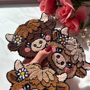 Op de afbeelding: Drie handgemaakte onderzetters in de vorm van een koe. Elke onderzetter heeft een bruin, beige en cr&egrave;mekleurig kleurenschema met een madeliefjes bloemenkrans. De onderzetters hebben roze wangen en blauwe ogen. De onderzetters zijn gemaakt van een textuur.
