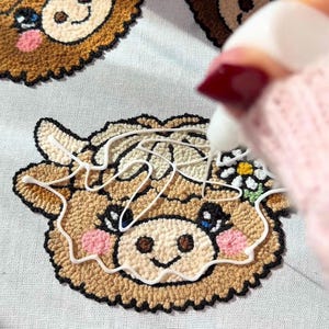 May include: Close-up of a craft project featuring three embroidered cow faces. The cow faces are brown with pink cheeks, black eyes, and a white flower. A hand with red nail polish is applying white glue to the cow face.