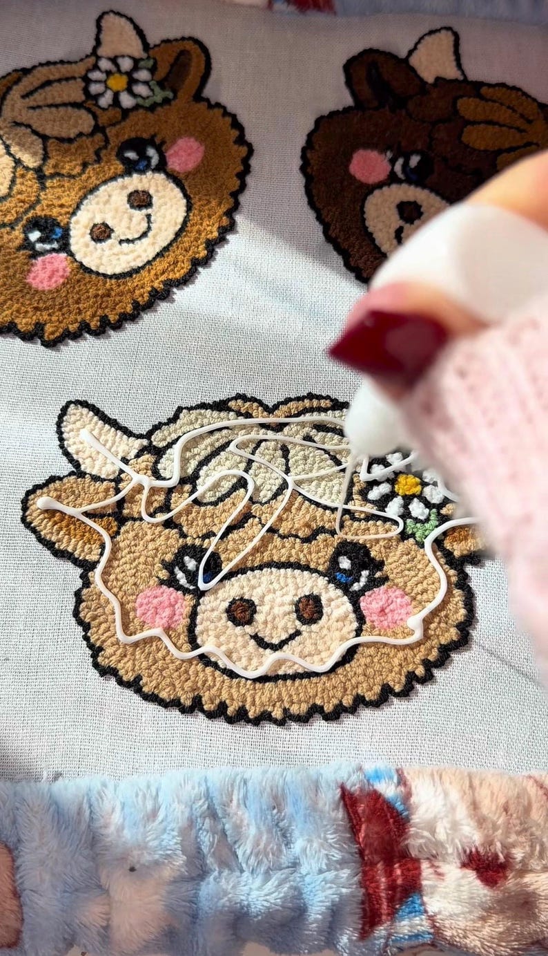 May include: Close-up of a craft project featuring three embroidered cow faces. The cow faces are brown with pink cheeks, black eyes, and a white flower. A hand with red nail polish is applying white glue to the cow face.