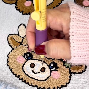 May include: A yellow and purple punch needle tool is used to create a brown, tan, and pink cow design on white fabric. The cow has a smiling face and blue eyes. The craft project is on a white surface with a blue and white fuzzy border.