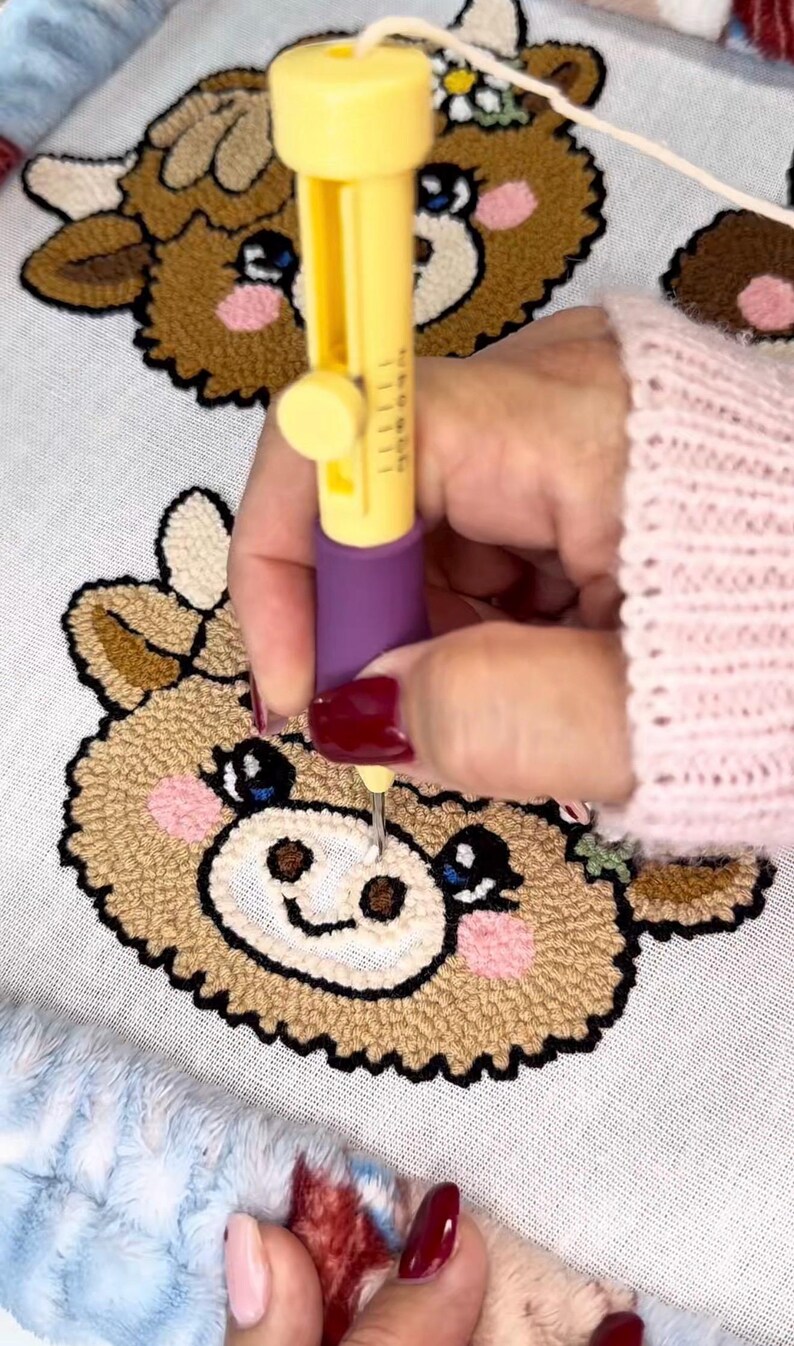 May include: A yellow and purple punch needle tool is used to create a brown, tan, and pink cow design on white fabric. The cow has a smiling face and blue eyes. The craft project is on a white surface with a blue and white fuzzy border.