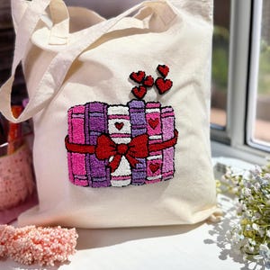 Può includere: Borsa tote color crema con un design ricamato colorato. Il design presenta una pila di libri nelle tonalità del rosa e del viola, legati con un fiocco rosso. Piccoli cuori rossi sono sopra i libri. La borsa ha due manici color crema.