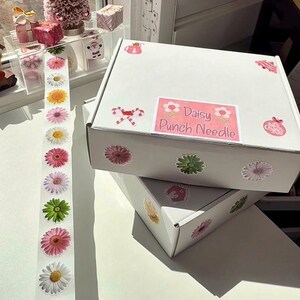 May include: Two white boxes adorned with daisy stickers and a label reading "Daisy Punch Needle." A strip of daisy stickers and a small pink Christmas tree are visible. The boxes are stacked on a white surface.